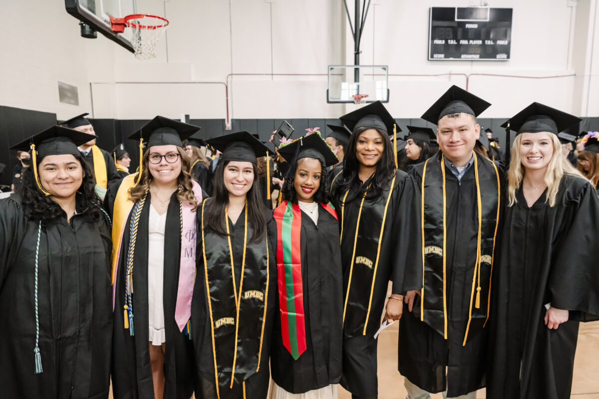 A group of seven graduates dressed in black caps and gowns stand together, smiling. They are indoors in a gymnasium, with basketball hoops visible in the background. Each graduate wears a stole or sash over their gown.