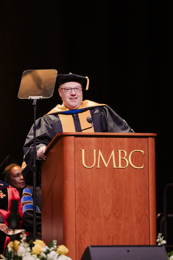 A man in academic regalia speaks at a podium marked UMBC during a graduation ceremony. The background is dimly lit, and there are people seated behind the speaker.