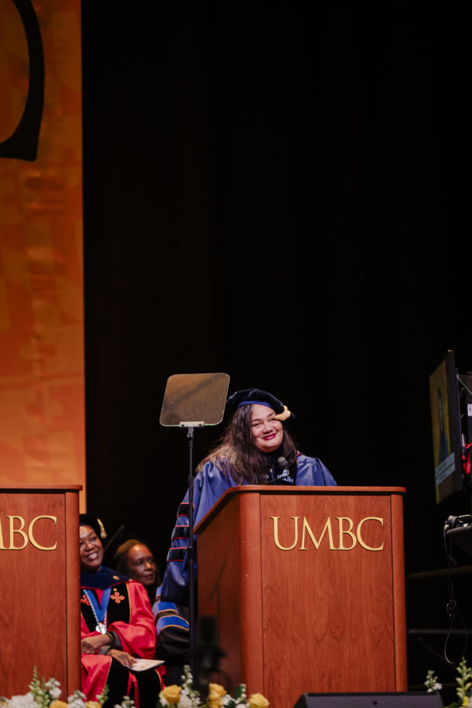 A woman in academic regalia speaks at a podium labeled UMBC during a graduation ceremony. Several people in caps and gowns are seated behind her. The background features a large orange banner with a faint pattern.