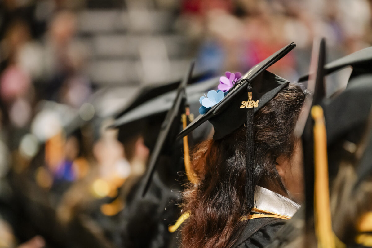 Rows of graduates in black caps and gowns sit in a ceremony. A cap in the foreground features the year 2024 and decorative flowers. The blurred background shows other graduates and attendees.