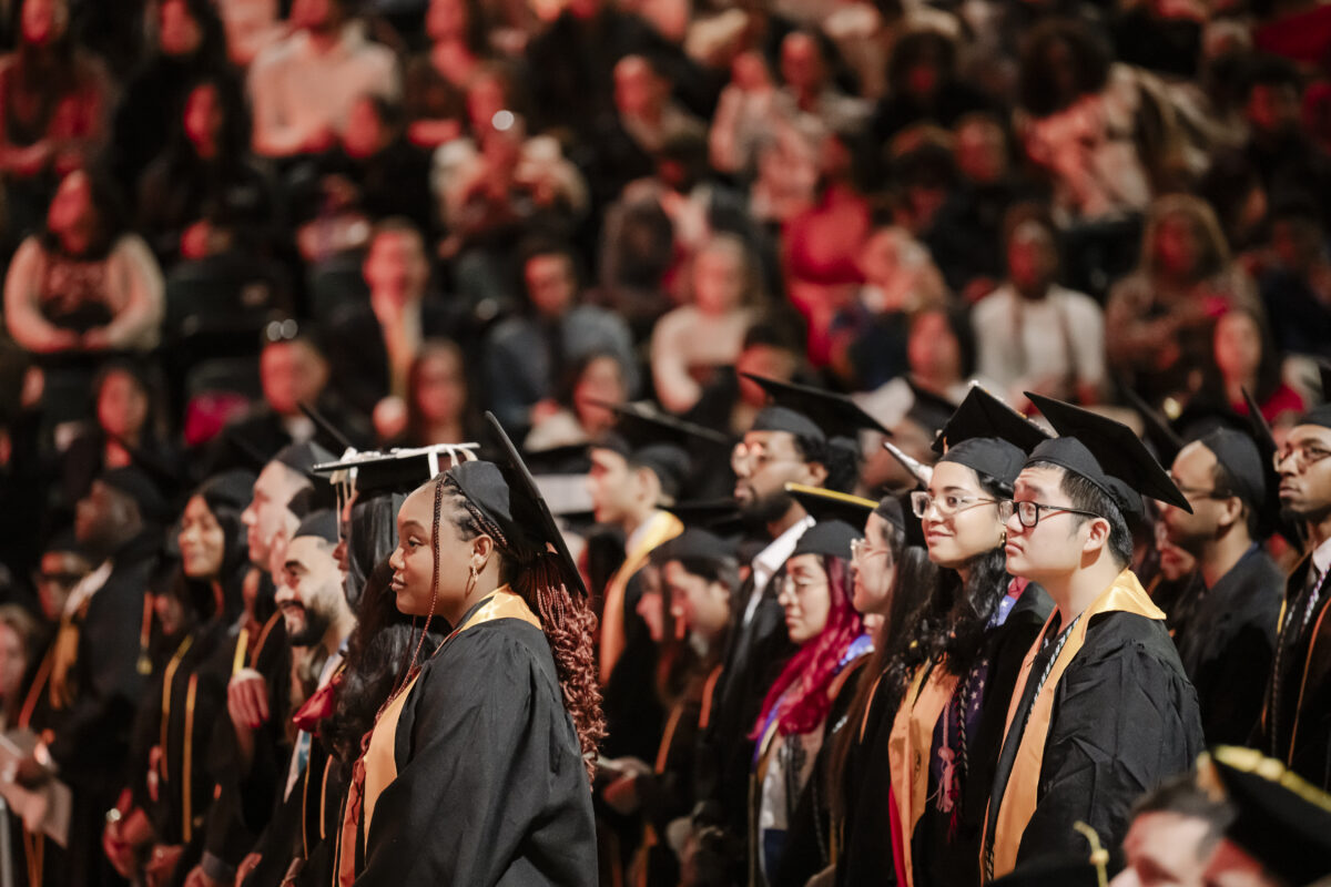 Graduates stand in rows wearing caps and gowns at a commencement ceremony. The audience is seated in the background, observing the event.