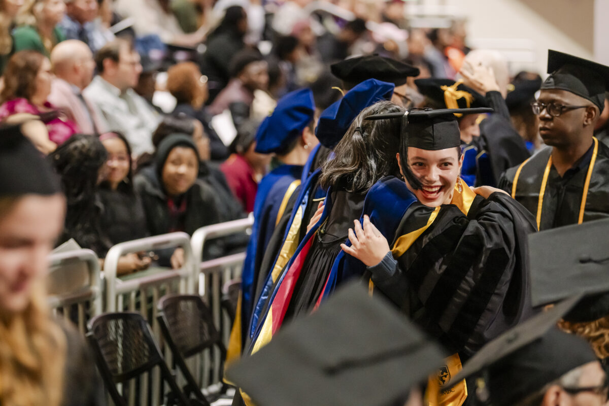 A joyful graduate wearing a cap and gown hugs another person during a graduation ceremony. Other graduates and attendees are visible in the background, seated and wearing academic regalia.