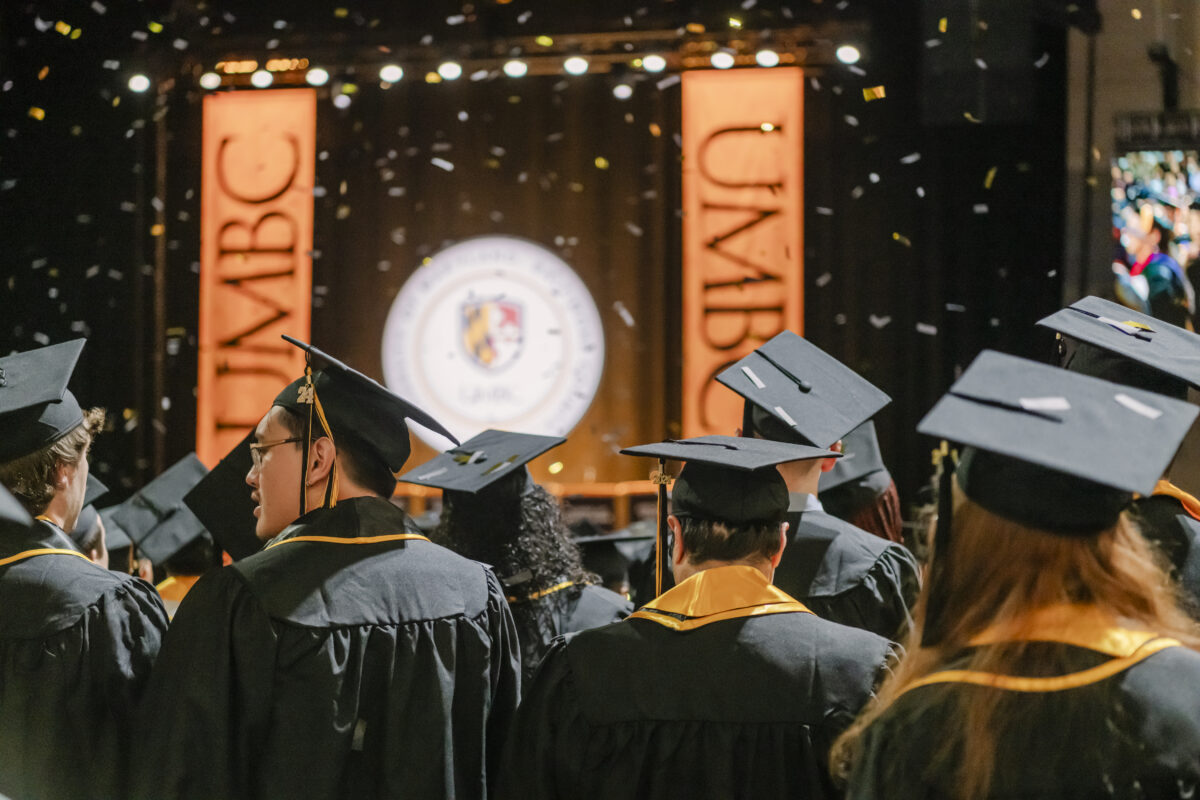 Graduates in caps and gowns celebrate at a commencement ceremony, with UMBC banners and a university seal in the background. Confetti is falling from above, adding a festive atmosphere.