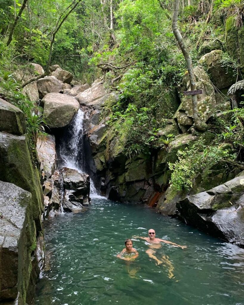 Two people swimming in a lake surrounded by boulders and greenery with a a waterfall in the background