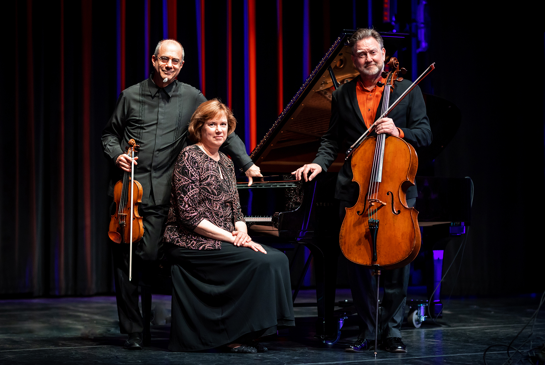 A trio of musicians pose on stage. A pianist in the middle, seated, is flanked on the left by a person holding a violin, and on the right a person holding a cello.