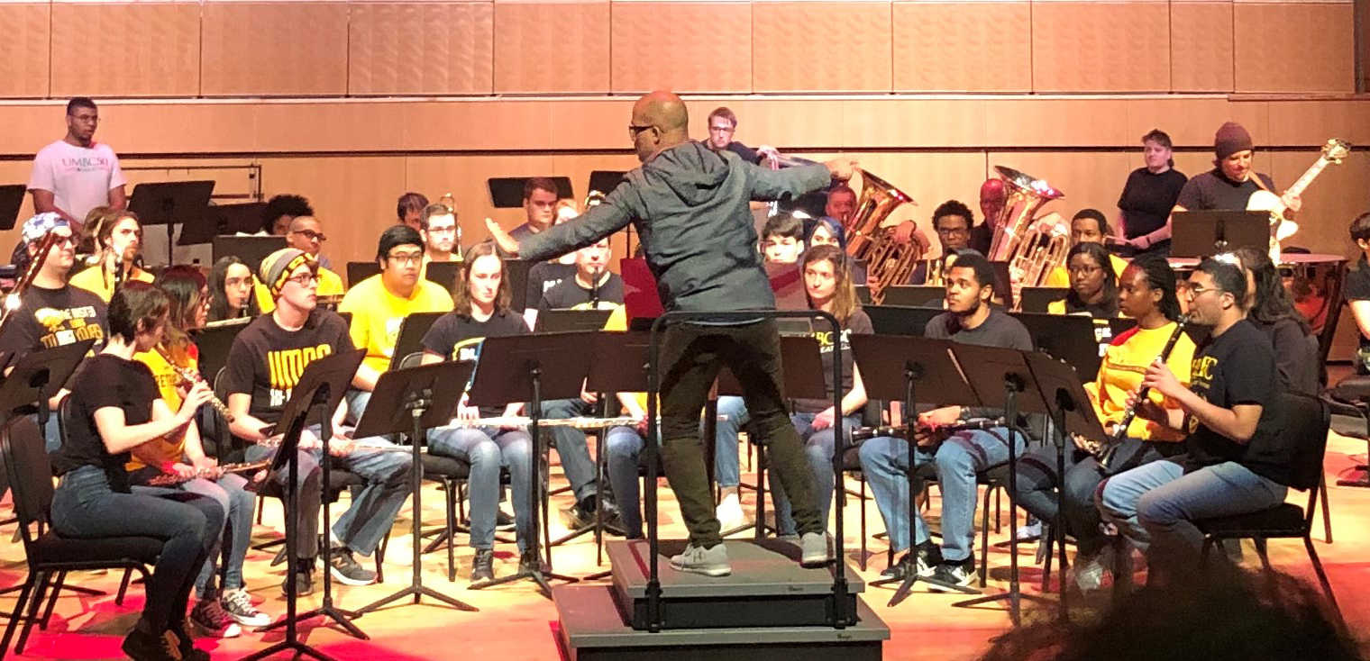 A person with a medium-dark skin tone, back to the camera, conducts an ensemble of musicians on a concert stage.