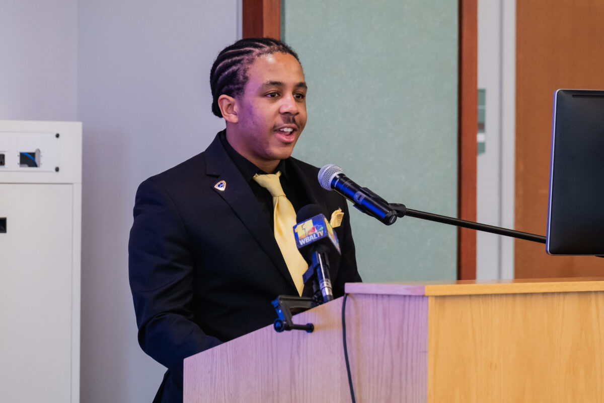 A college student wearing a black suit and gold tie stands at a podium speaking into a microphone about Health and Human Services.