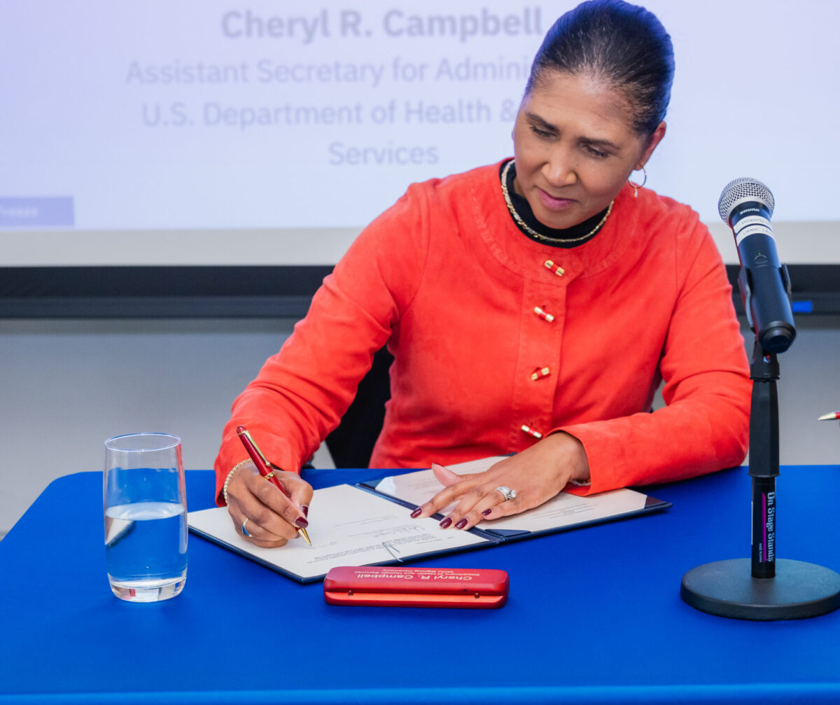 A business person sitting at a conference table signing a legal document with a projection screen in the background.