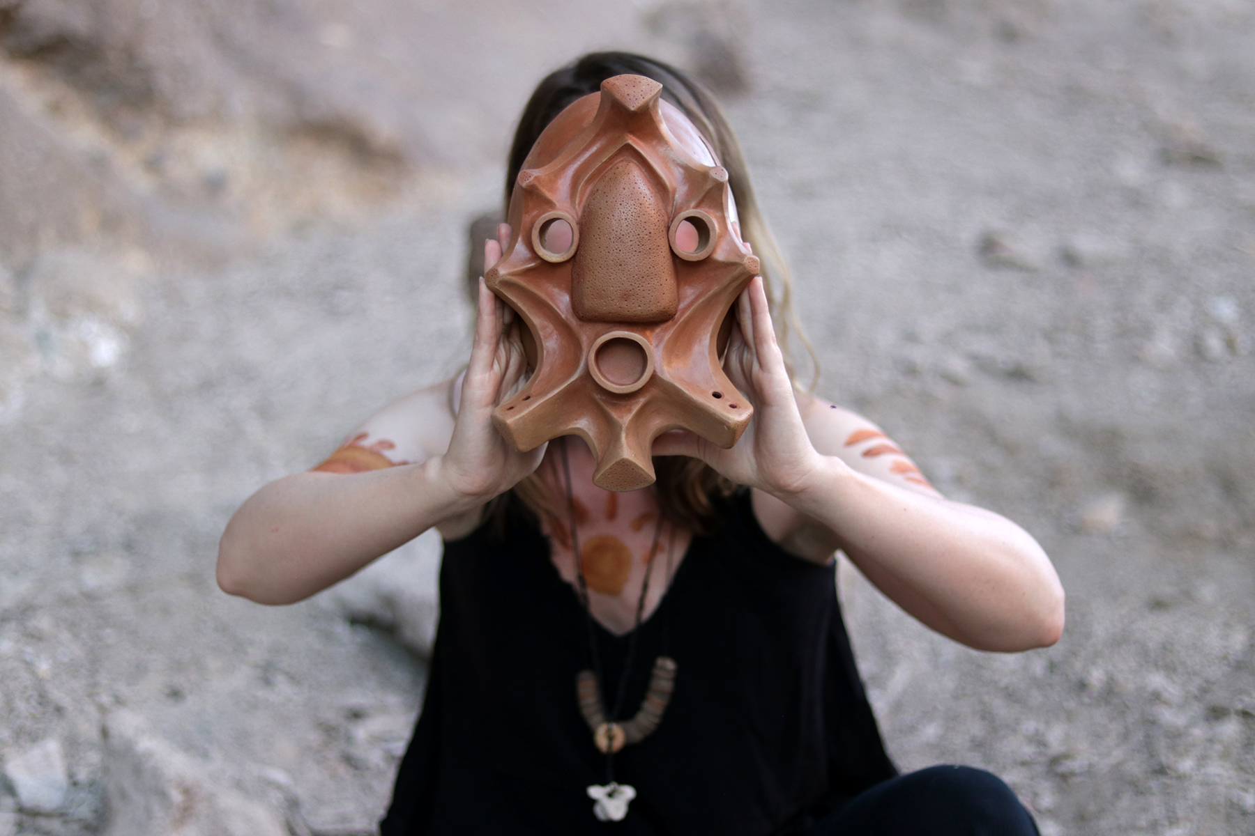 A person wearing a dark outfit holds a clay mask over their face.