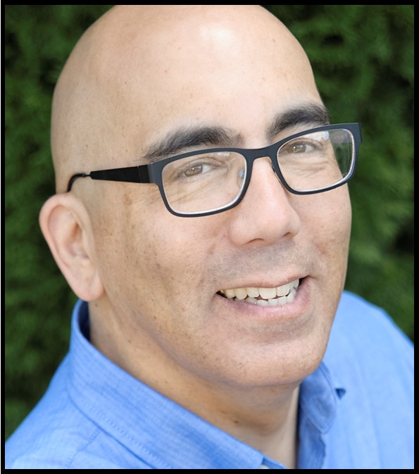 A person with a medium-light skin tone, bare head and glasses, wearing a blue shirt, smiles at the camera.