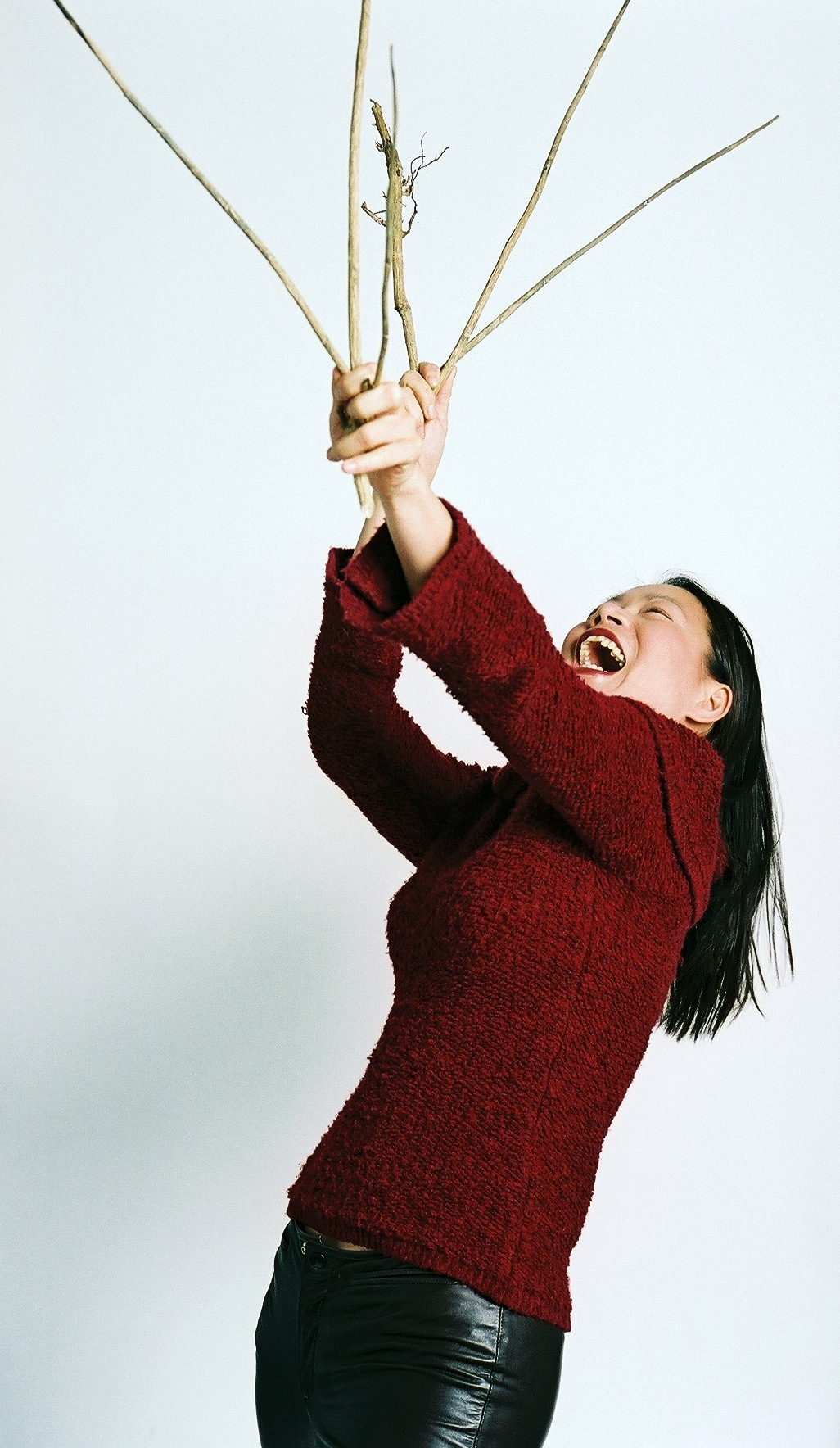 A person with a medium skin tone, wearing a red sweater and black pants, laughs as they look up at a cluster of sticks in their hands.