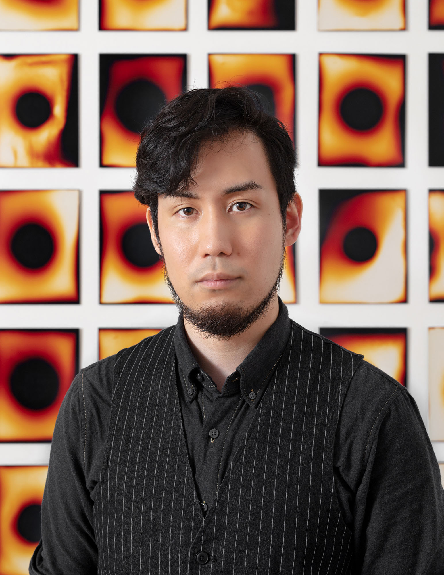 A person with a medium skin tone, black hair and beard, wearing a black shirt and vest, poses in front of a rectangular grouping of black and orange images.