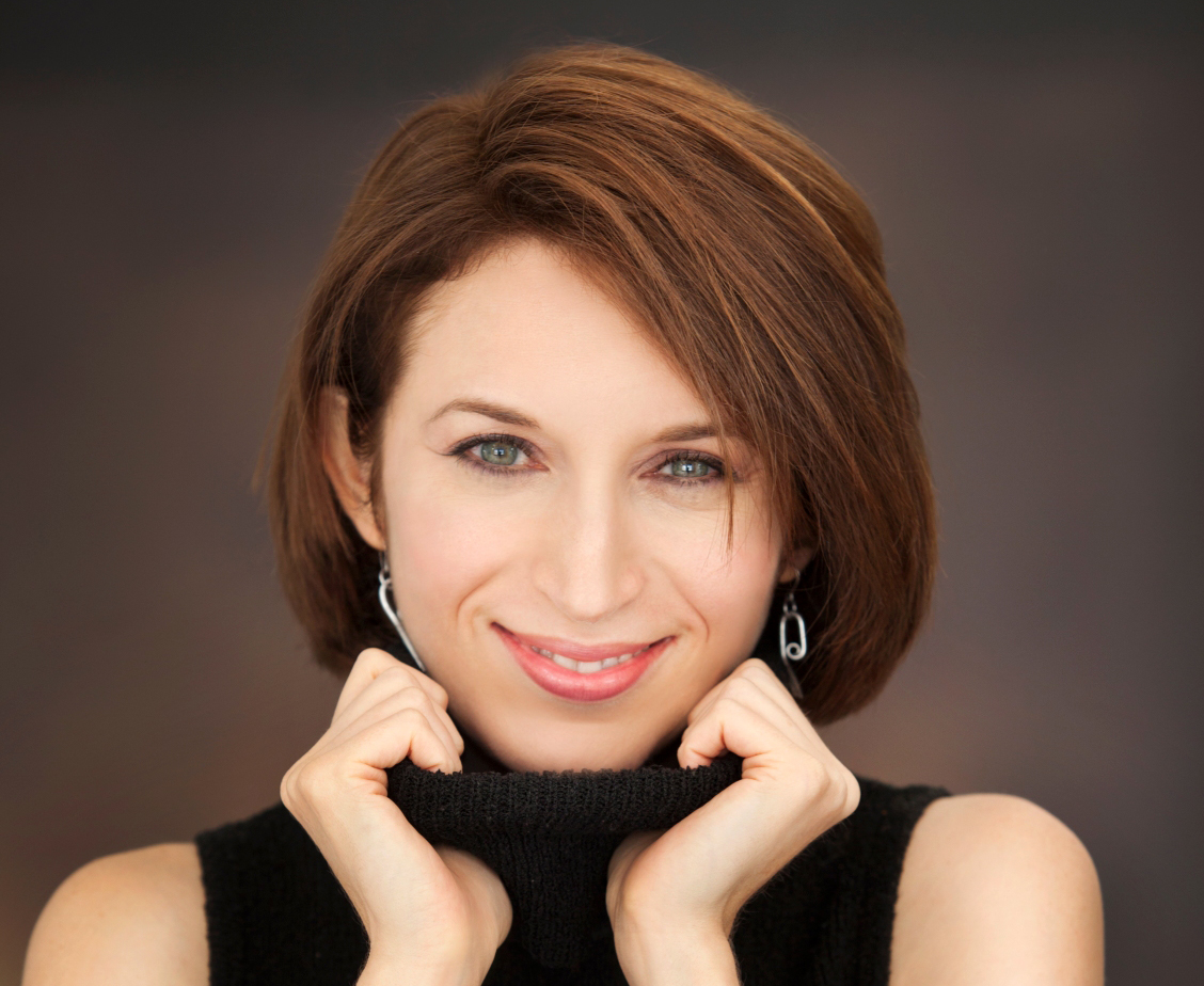 A person with a light skin tone, brown hair and blue eyes, smiles at the camera while pulling a turtleneck up toward their chin.