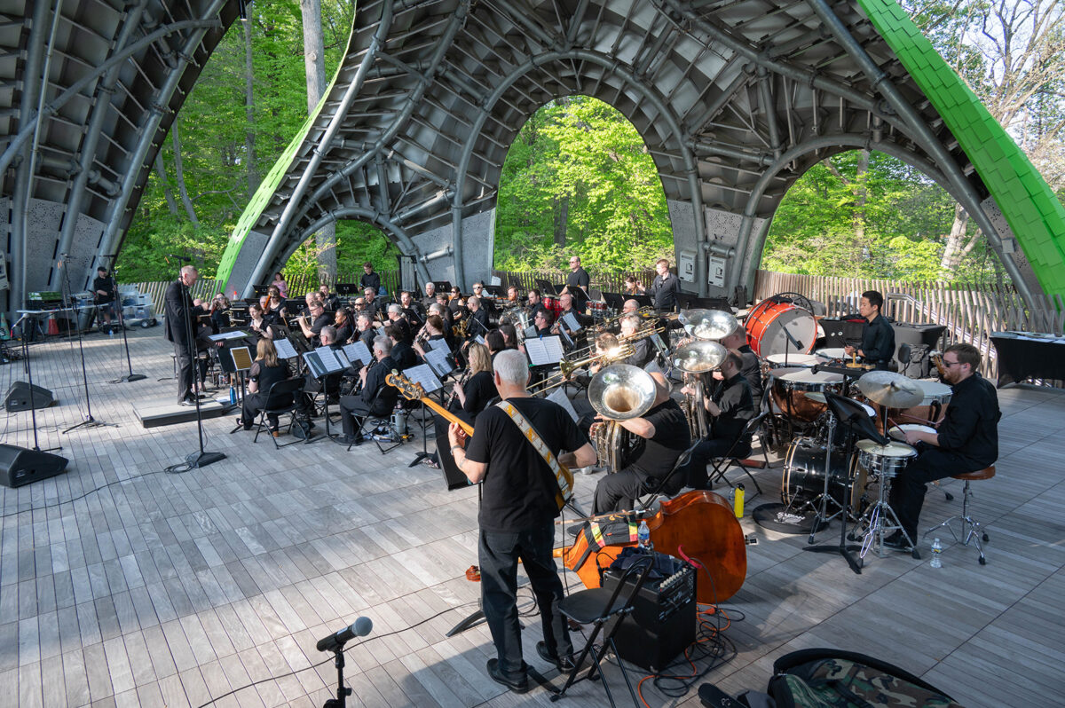 An ensemble of about 40 musicians perform under massive arches in an outdoor setting.