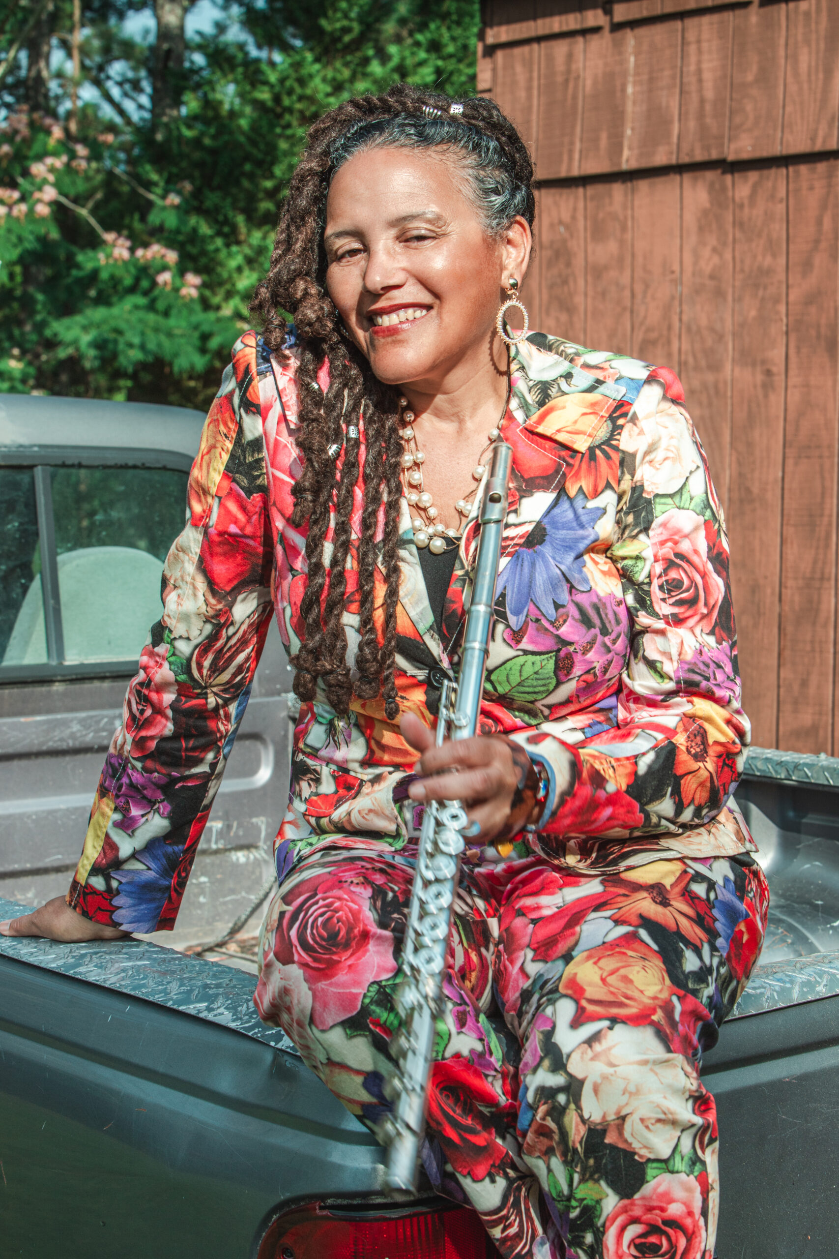 A portrait of flutist Nicole Mitchell. A Black woman, she wears a colorful outfit, holds a flute, and sits on the hood of a truck.