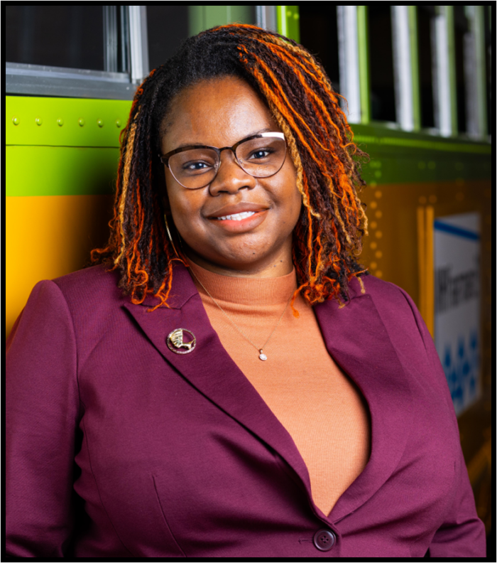 A person with a medium dark skin tone, wearing glasses and a purple jacket, smiles at the camera. Her dark braided hair has orange highlights.