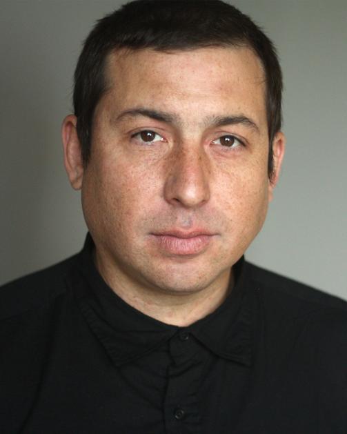 A portrait of Tommy Orange, an indigenous man, wearing a black button-up shirt. He has short dark hair.