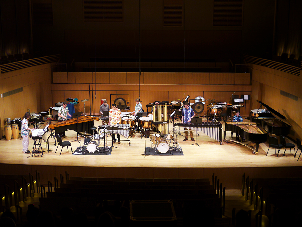 A group of musicians perform on percussion instruments, on a concert stage filled with instruments.