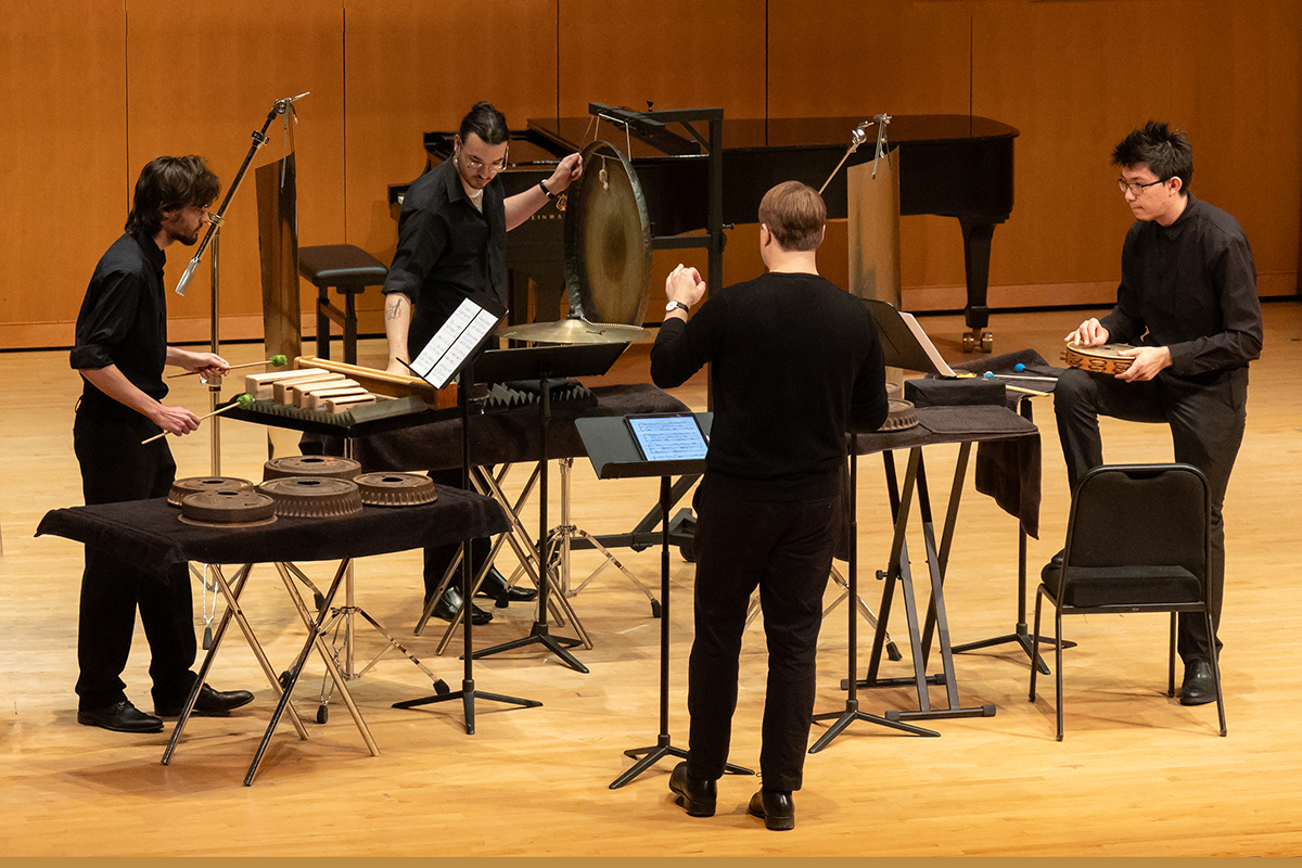 Four percussionists perform on a concert stage with a conductor.