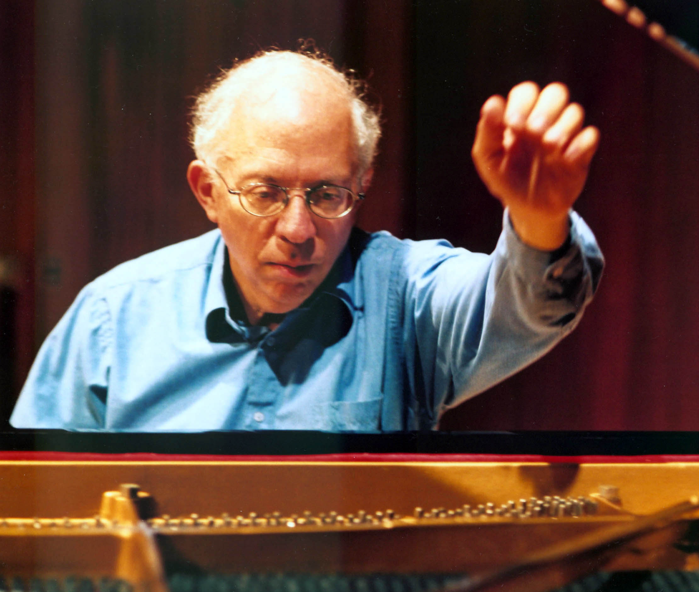 Seated at a grand piano, and viewed from the perspective of the strings, a person with glasses and a light blue shirt raises their left hand, as if about to play.