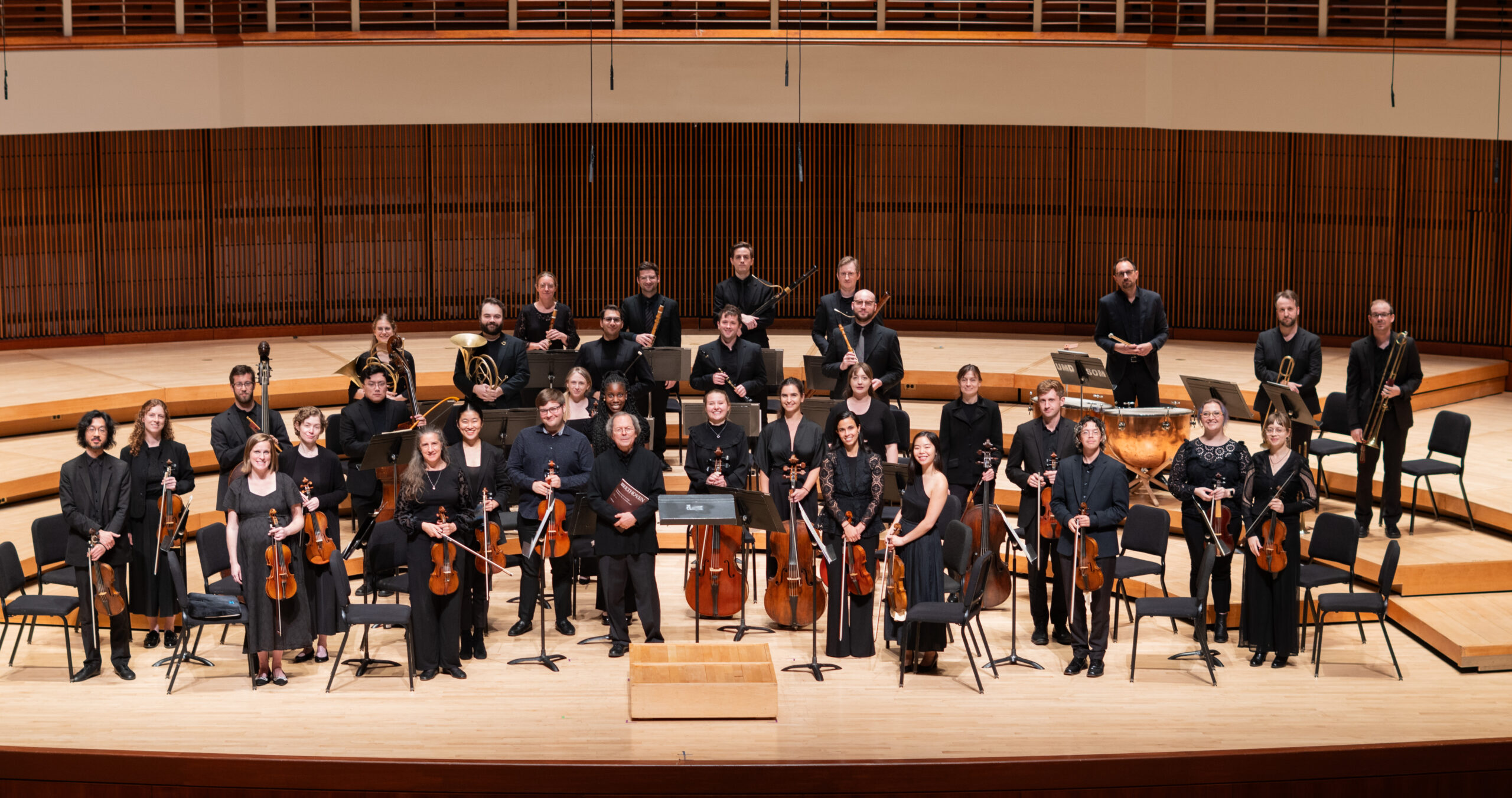 A classical orchestra of 34 people stands on a concert stage.