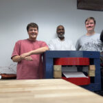 Four people pose in lab near structure with large metal components (magnet).