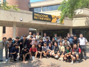 A large group of Japanese high school students gather under a walkway on a paved path on a summer day
