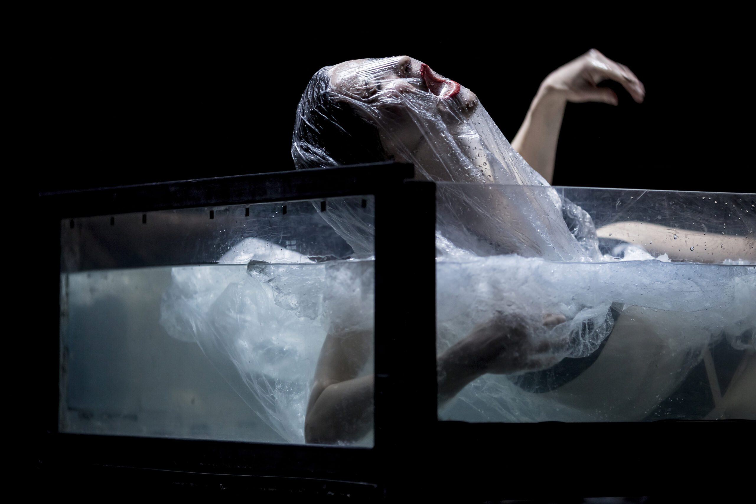 A person, wrapped in plastic, gapes with an open mouth, half submerged in a glass tub of water.