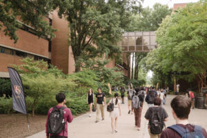 Students walk along pathway between brick academic buildings, trees on either side.