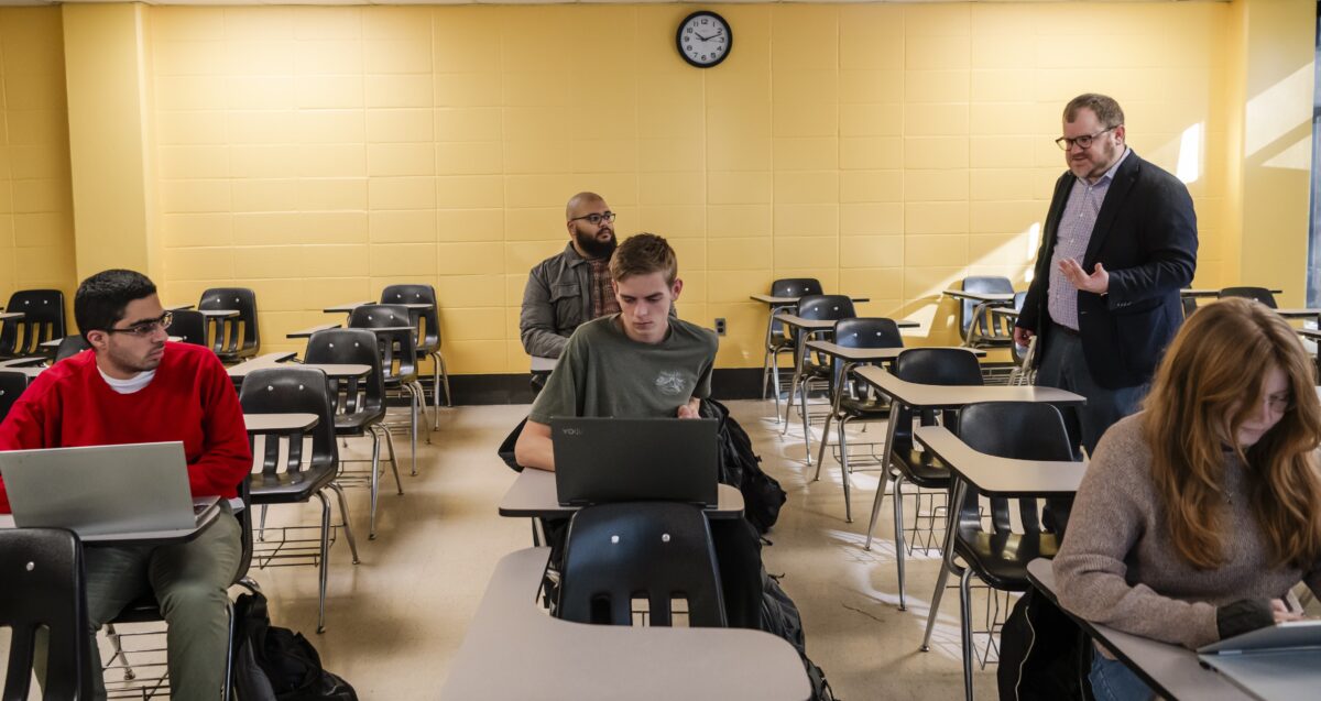 A college professor walks between student desks as he lectures in a classroom with a yellow brick wall in the background