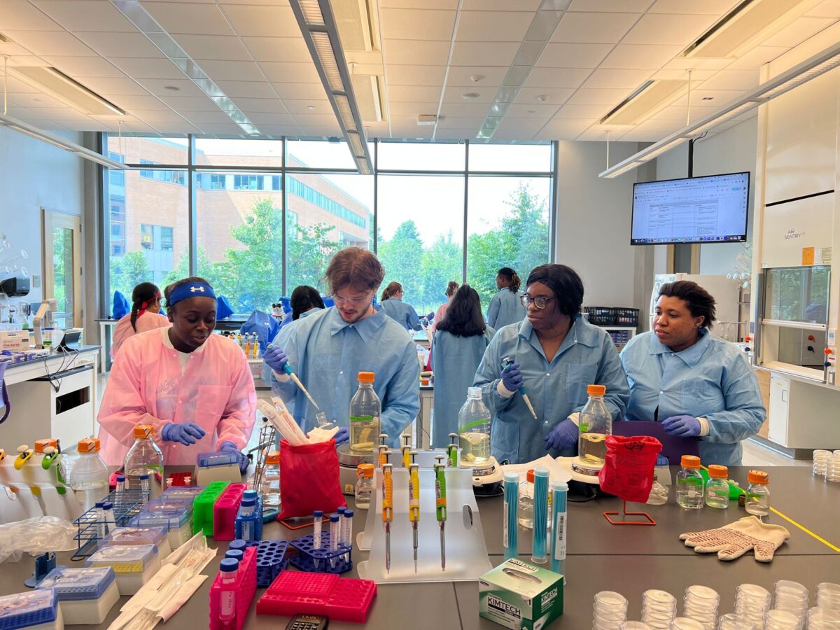 four students in lab coats and safety goggles working at a lab bench with pipettes, flasks, and bacterial plates