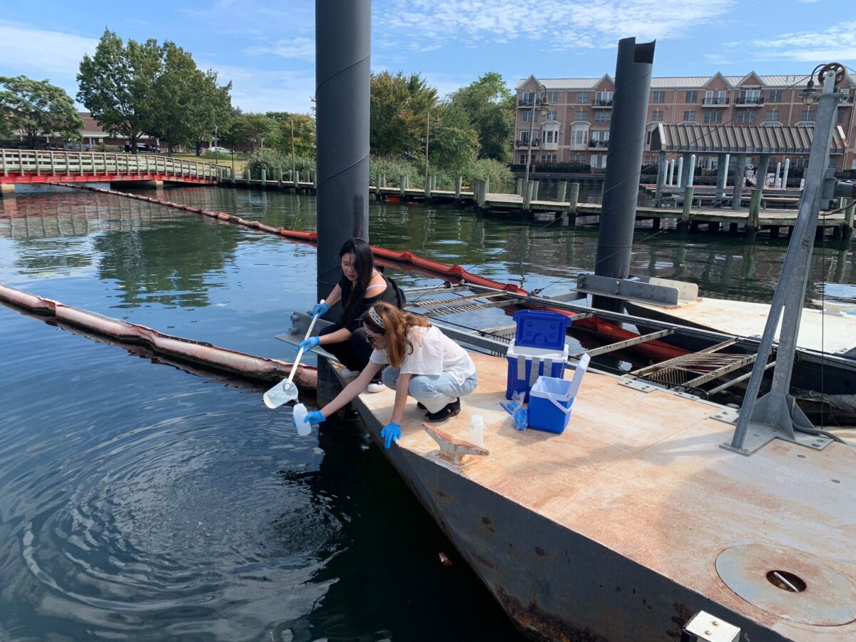 Two people crouch on a pier near the water. One person pours water from one container into a bottle that the other person holds.
