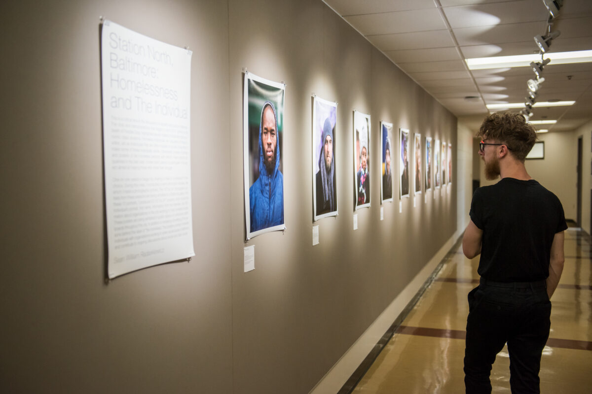 A college student looking at a series of images hung up on a wall in a hallway. There is a placard next to the images on the left hand side that says "Station North, Baltimore Homelessness and The Individual."