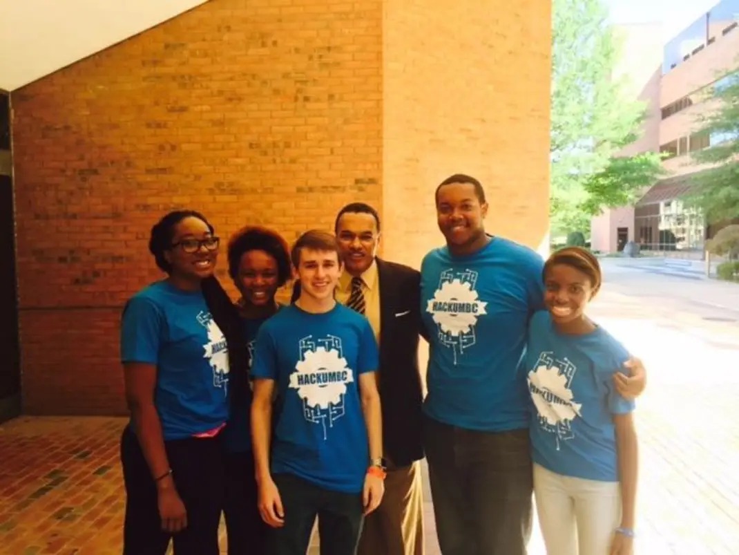 Group of young people wearing blue shirts that read "HACKUMBC" pose with man in suit and tie.