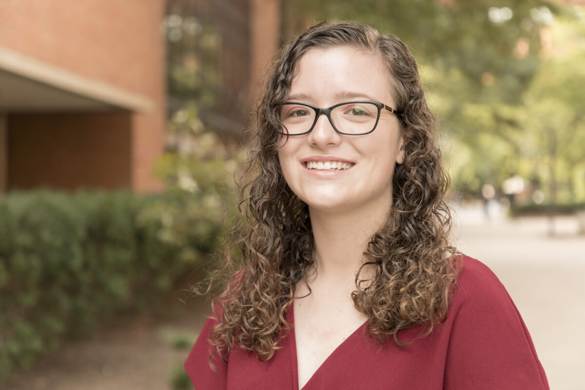 A college student with long, curly, brown hair with dark rimmed glasses stands outside in front of red brick buildings
