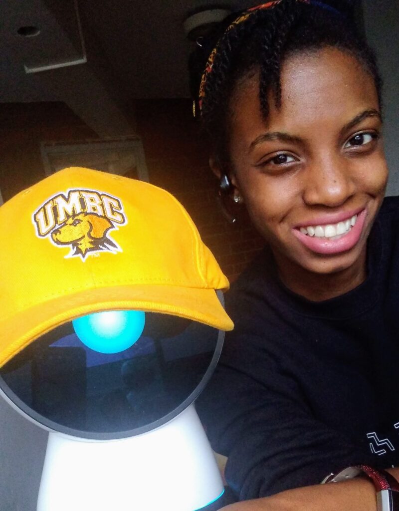 A young woman next to a robot. The robot wears a yellow hat that reads "UMBC"