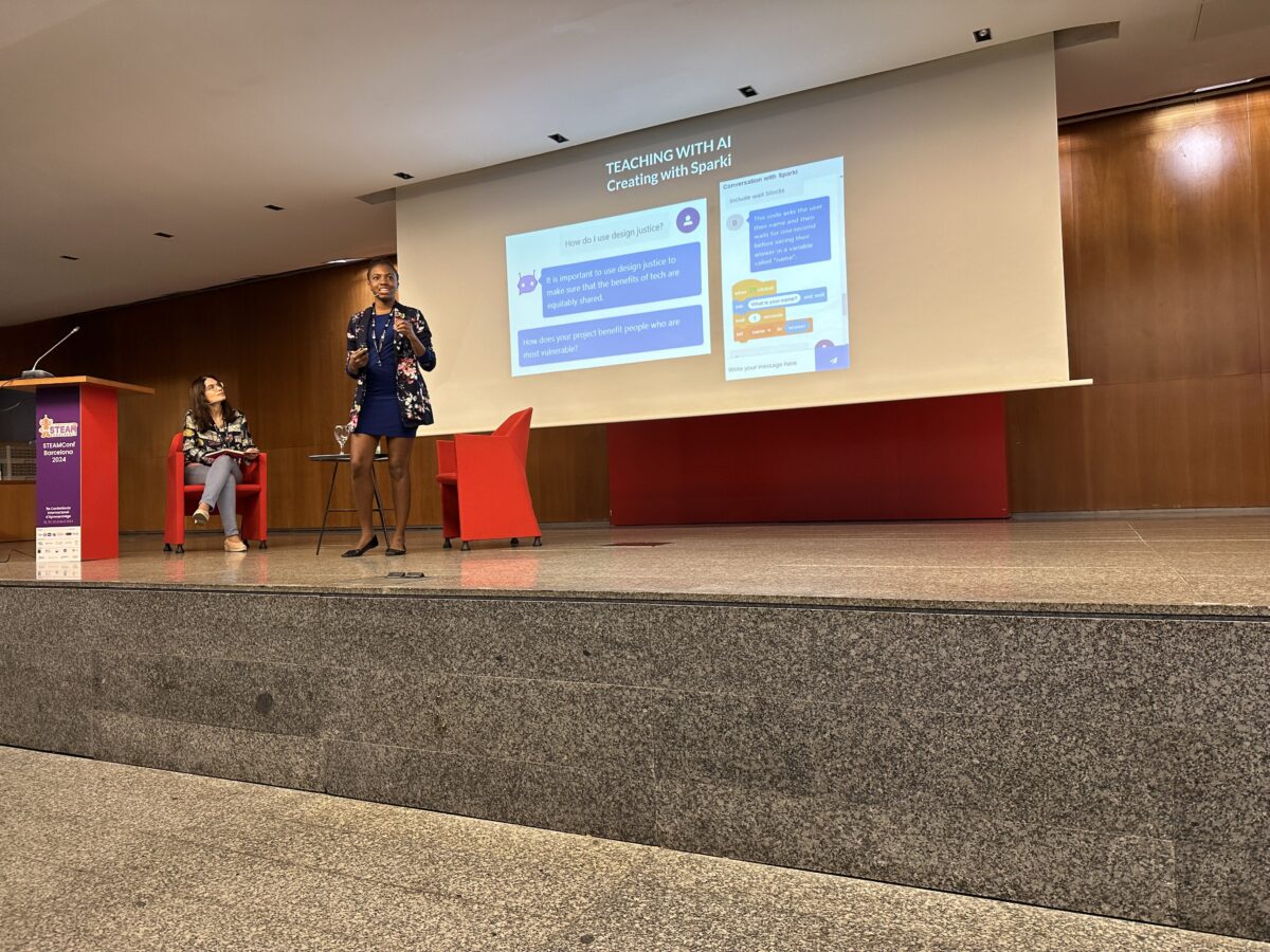 Woman stands on stage in front of presentation screen, holding microphone.