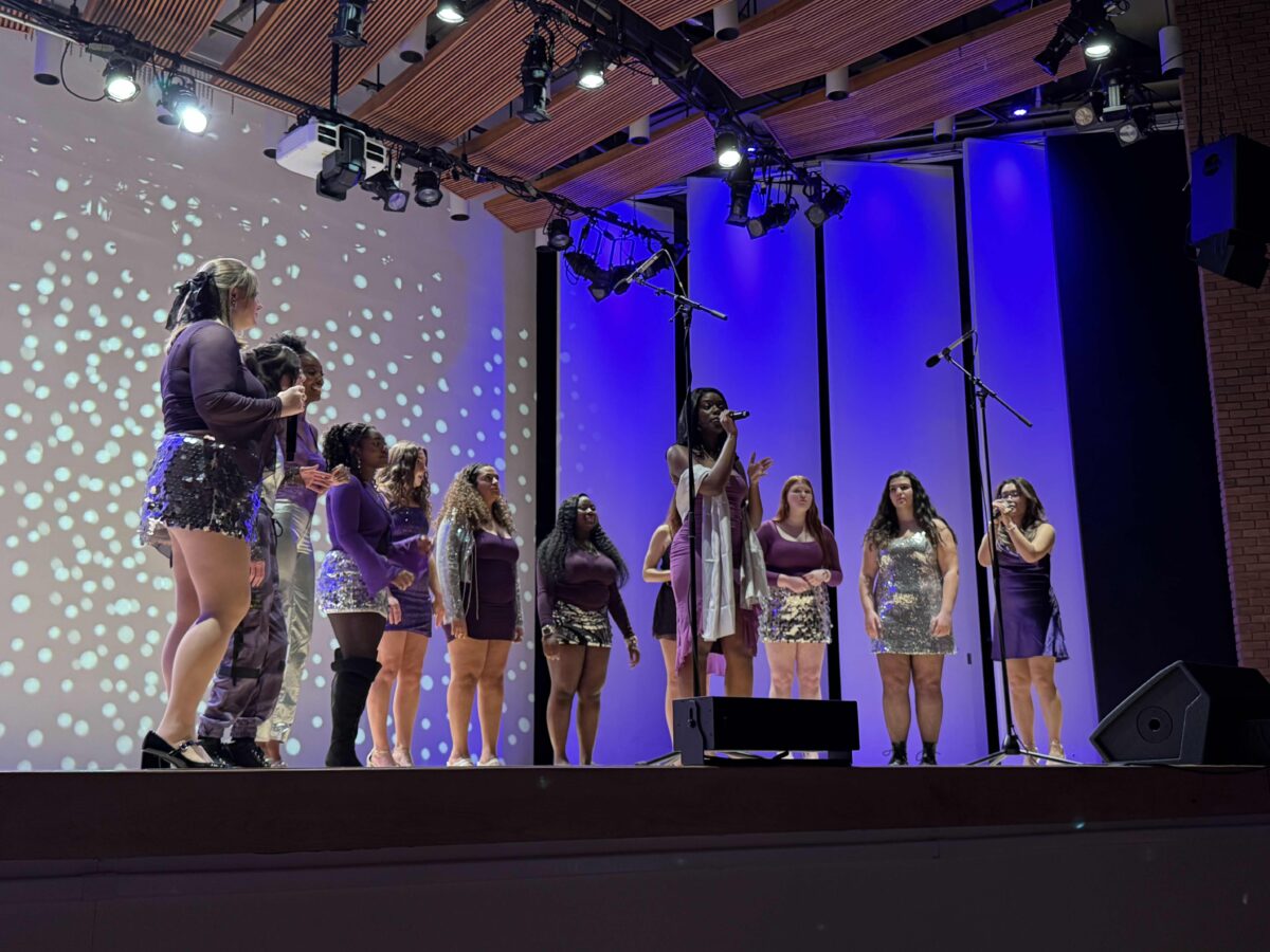 women in purple and silver outfits standing in a semicircle on stage; one holds a microphone in front