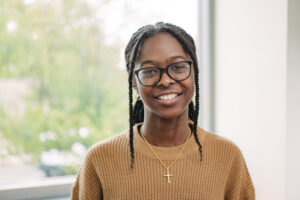 A young woman, Dionne Cole, with glasses smiles for a headshot, wearing a tan sweater and a cross around her neck.