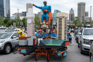 Kinetic sculpture with teal man in wheelchair and cape, pedaled by team wearing blue shirts