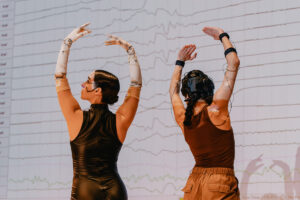 Two dancers perform in front of screen displaying wavy line signals.