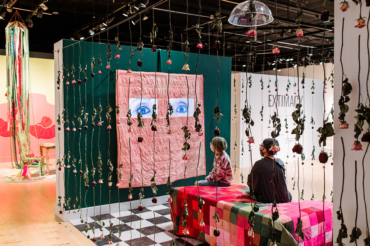 Arts exhibit at UMBC's CADVC gallery, featuring strings of roses hanging from the ceiling, and a large pink piece with painted eyes