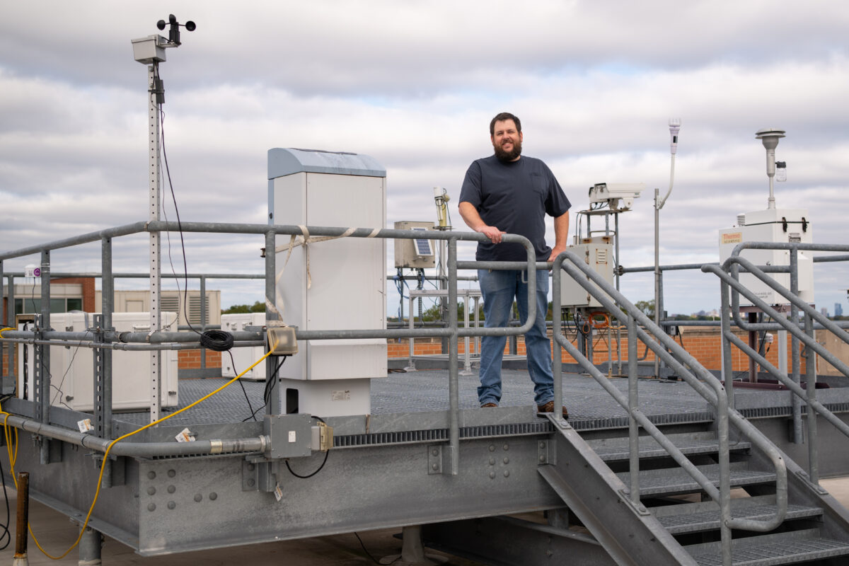 man stands on metal paltform on the roof of a building; several large scientific instruments sit behind him on the platform