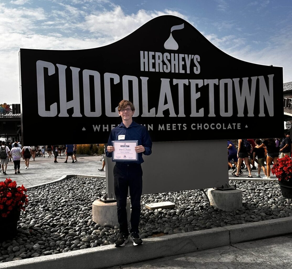 A young man stands in front of a Hershey's Chocolatetown sign holding a certificate of achievement for his summer internship