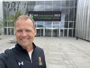 A person smiling in front of a modern building with an electronic sign reading "HCI International 2023, 23-28 July, Copenhagen, Denmark."