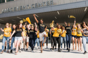 a large group of students wearing black and gold spirit wear, jumping and cheering in front of the UMBC event center