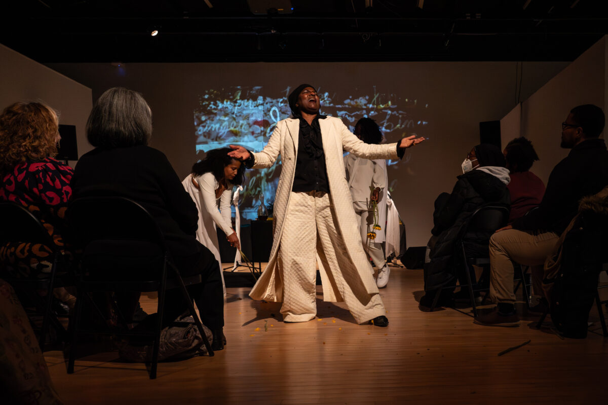Before a seated audience, a woman spreads her arms apart in a theatrical gesture.