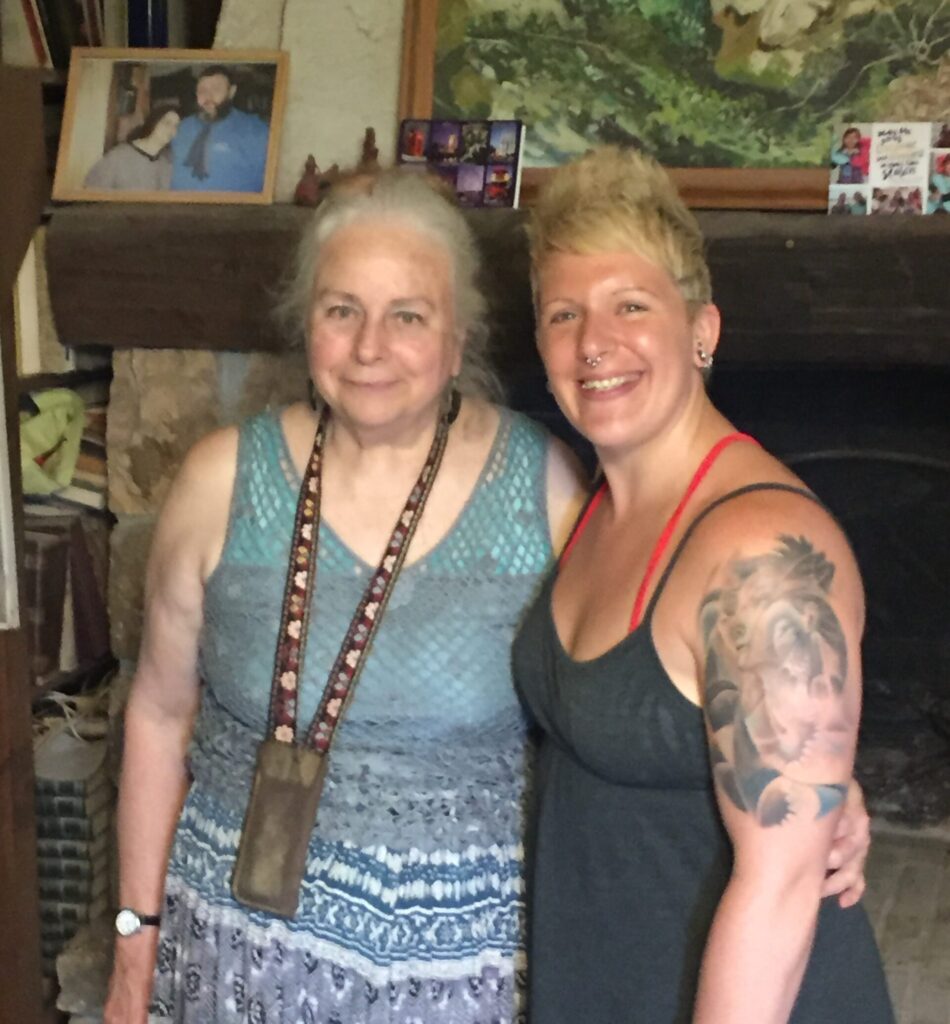 One woman wearing a blue dress stands next to another woman with a black tank top and a large tattoo on her arm. erotic
