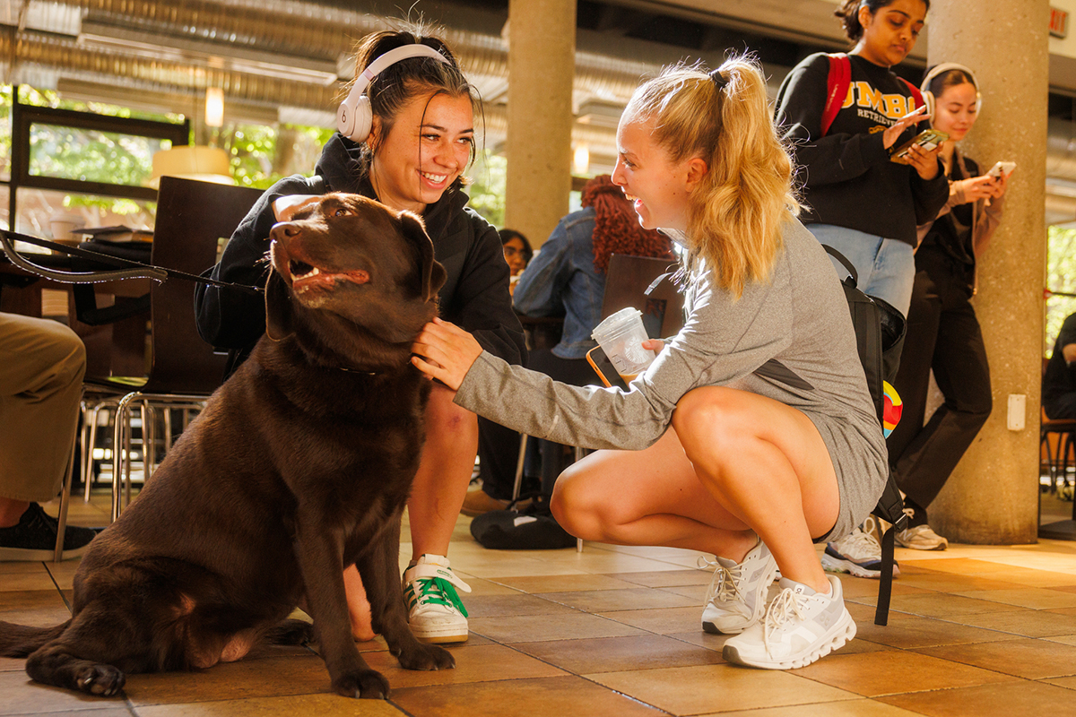 two young women petting UMBC's mascot dog, Chip the black labrador retriever