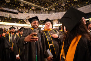 Two students post for a selfie throwing a peace sign while confetti falls at UMBC's graduation